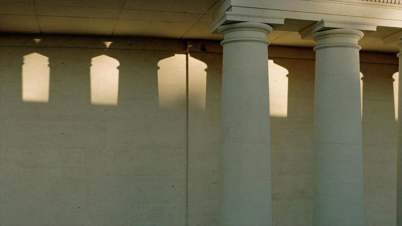 A limestone colonnade in late afternoon light, casting long geometric shadows across a pale wall.