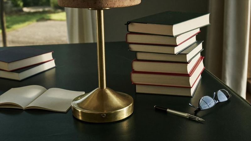 A dark green leather desk with a brass lamp, a stack of clothbound books, an open notebook, a pen and reading glasses.